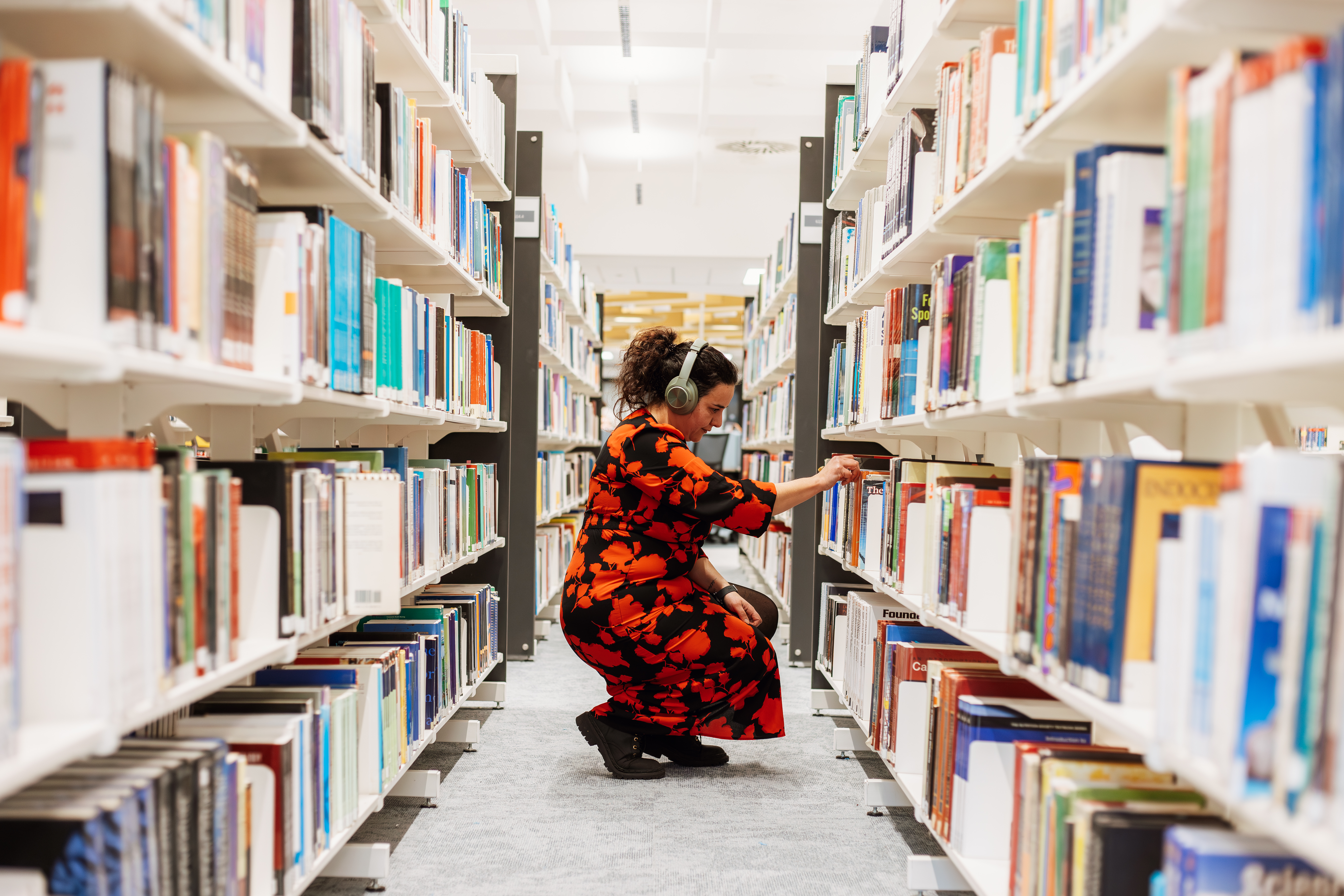 Student wearing headphones and a bright coloured dress crouched between rows of books, looking at a book on a shelf.