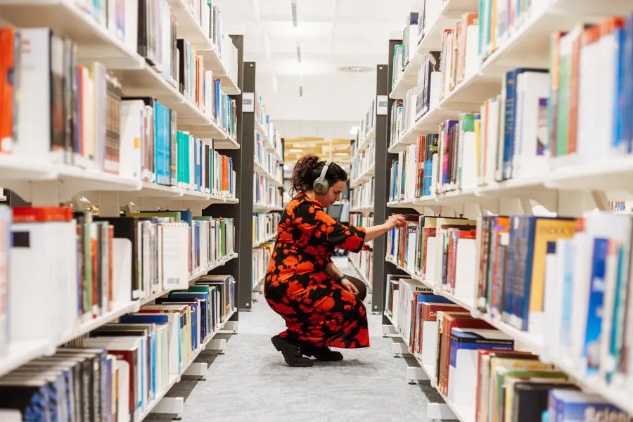Student wearing headphones and a bright coloured dress crouched between rows of books, looking at a book on a shelf.