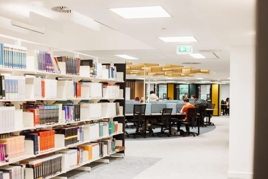 Shelves of books to the left with study spaces and students studying on the right.